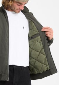 Militarna zielona kurtka bomberka z pikowaną podszewką; ma zamek błyskawiczny z przodu, ściągacze na rękawach i biały T-shirt z małym logo.