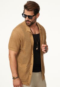 Strickjacke mit kurzen Ärmeln in Hellbraun, offenem Netzdesign, Brusttasche und Knopfverschluss. Getragen über einem schwarzen Tanktop.