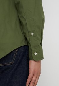 Olivově zelená košile s dlouhým rukávem a manžetami na knoflíky, s přizpůsobeným střihem, bavlněnou látkou a kontrastním švem. Nošena s tmavými džínami.