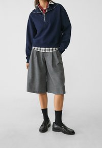 Marineblauwe half-ritssweatshirt over een grijze geruite broek met plooien, gecombineerd met zwarte loafers en enkelsokken, voor een gelaagde look.