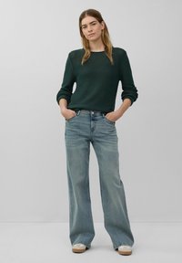 Dunkelgrüner strukturierter Pullover kombiniert mit hellblauen weit geschnittenen Jeans. Die Jeans sind leicht ausgebleicht und haben ein klassisches Fünf-Taschen-Design.