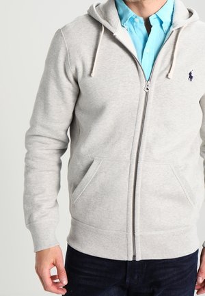 Man wearing a light gray zip-up hoodie over a blue collared shirt and dark pants, standing against a plain background.