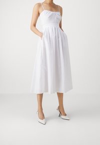 Vit midiklänning med tunna axelremmar, texturerad tyg, samlad byst och fickor; kombinerad med spetsiga vita klackar.