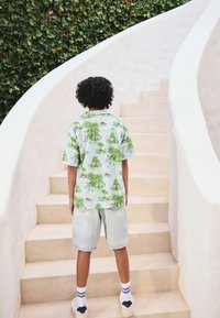 Chemise à manches courtes avec un fond bleu clair, ornée d'un motif tropical vert. Portée avec un short en denim clair et des baskets blanches.