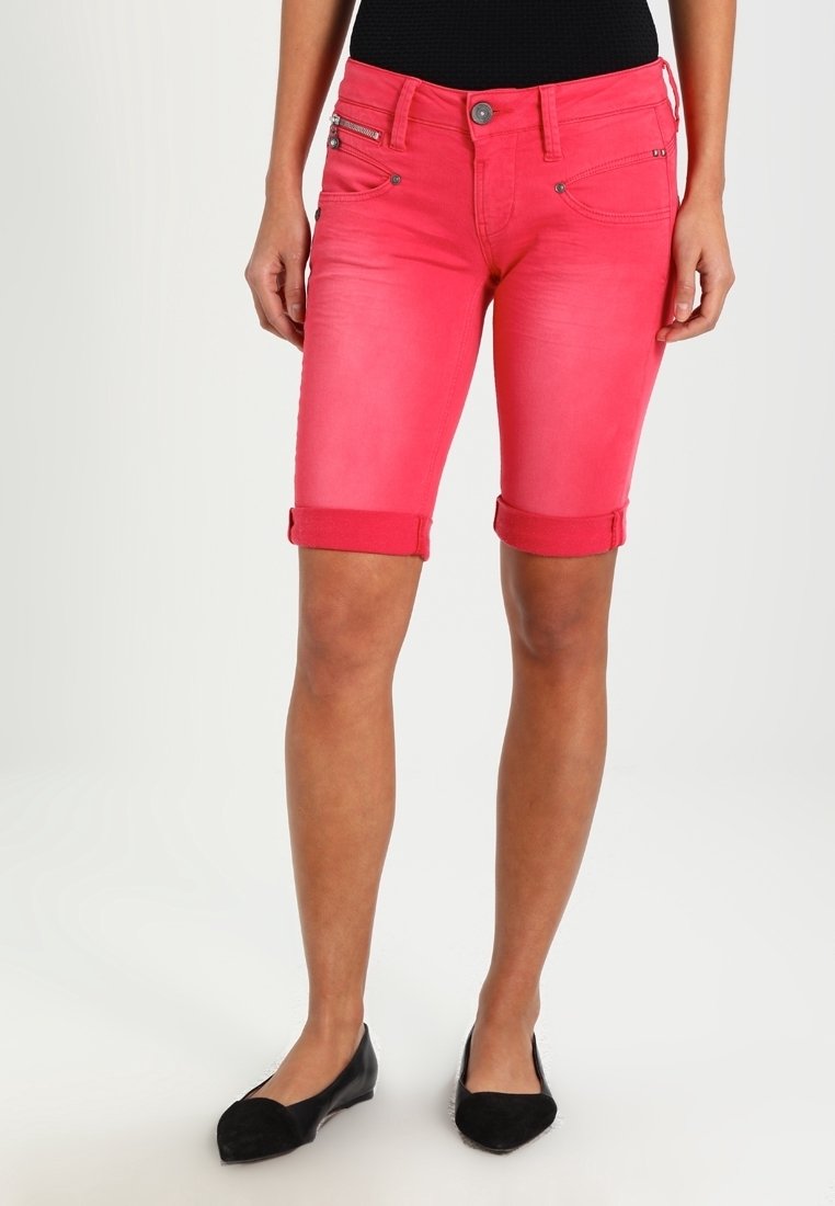 Femme portant un short rouge vif remonté jusqu'aux genoux et des chaussures plates noires, debout devant un fond clair uni.