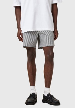 Randiga shorts i svart och vitt tyg, med ett elastiskt midjeband; bärs med en vit t-shirt och svarta skor med vita strumpor.