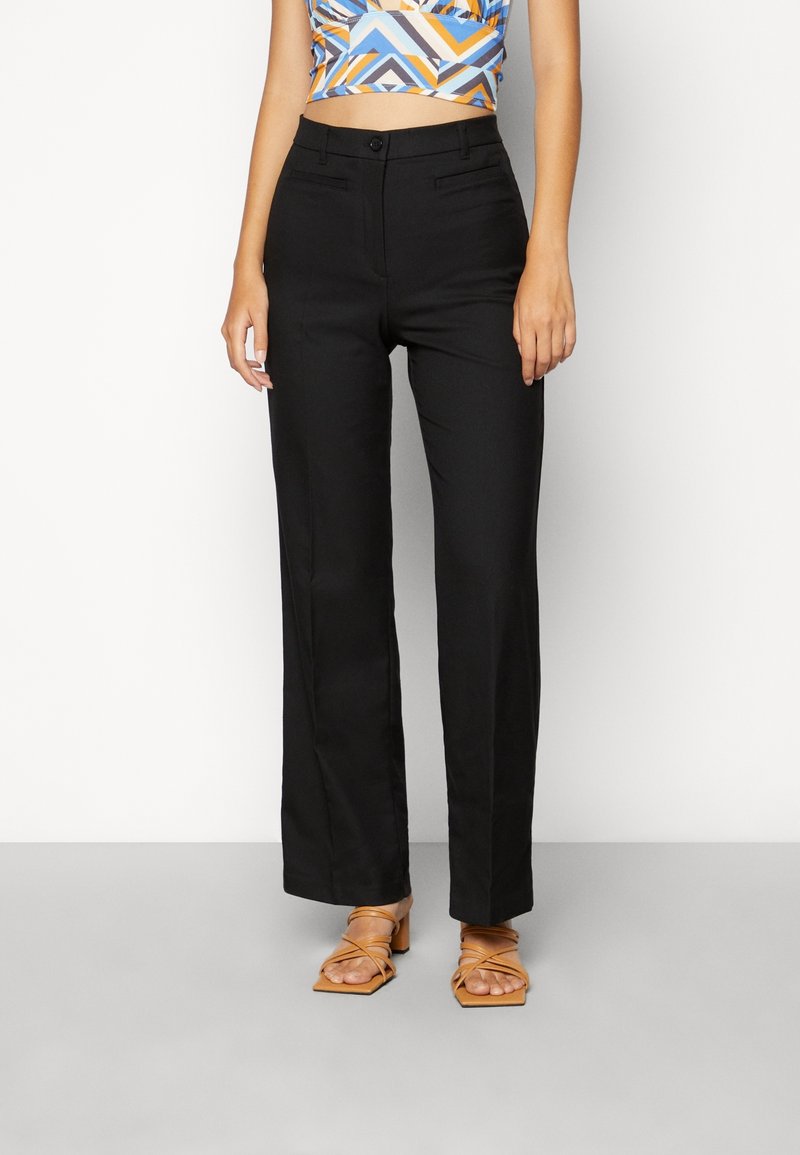 Femme portant un pantalon noir droit, un crop top géométrique coloré et des sandales à talons blocs beige à brides, debout sur un fond blanc.