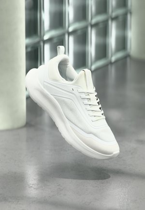 Sneaker bianca Calvin Klein con dettagli in mesh e lacci, che sembra galleggiare sopra un pavimento in cemento accanto a un muro di blocchi di vetro.