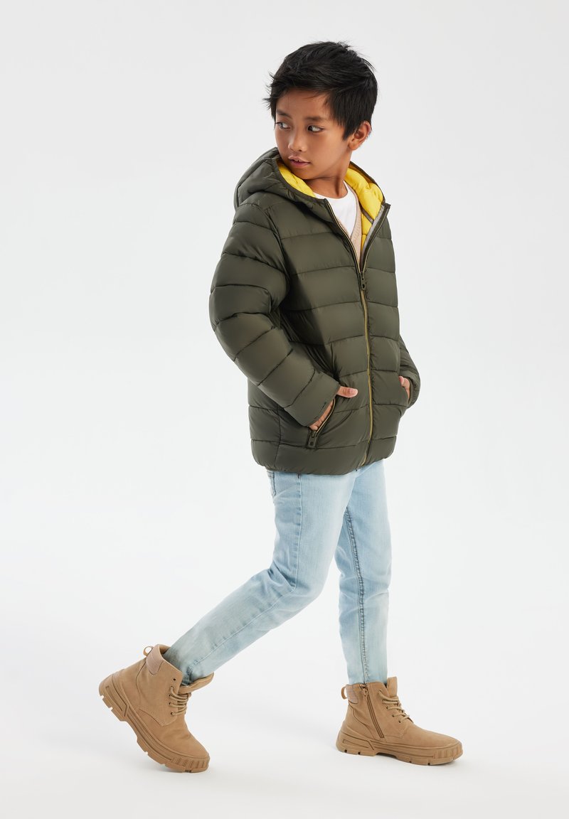 Olive green padded jacket with hood, yellow lining, front zipper, side pockets. Paired with light blue jeans and tan ankle boots.