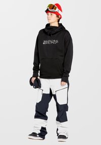 Schwarzer Hoodie mit "Volcom Snow"-Logo, kombiniert mit weiten weißen und marineblauen Schneehosen mit Reißverschlüssen. Zubehör umfasst eine rote Mütze und eine Skibrille.