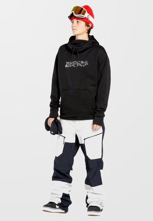 Zwarte hoodie met het "Volcom Snow" logo, gecombineerd met baggy witte en marineblauwe sneeuwbroeken met ritsen. Accessoires omvatten een rode beanie en een skibril.