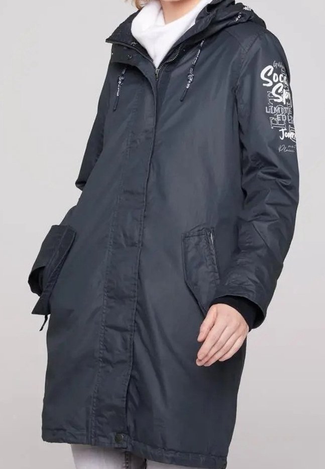 Kobieta ubrana w czarną parkę z kapturem, z zamykanymi kieszeniami, białym szalikiem oraz nadrukowanym tekstem na górnej lewej rękawie.