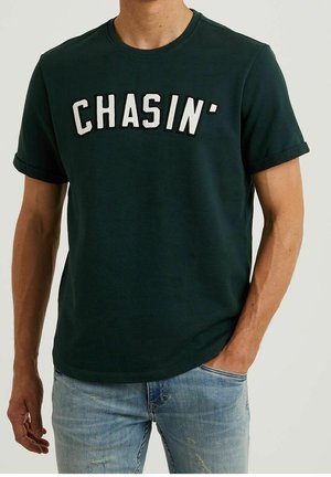 Homme portant un t-shirt vert foncé à manches retroussées avec des lettres blanches formant le mot « CHASIN' », associé à un jean bleu clair déchiré.