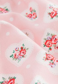 Tissu rose tendre avec des formes de cœur blanches contenant des grappes de roses rouges, roses et violettes et des feuilles vertes, drapé de plis doux.