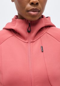 Κοντινό πλάνο ενός ατόμου που φοράει μια ροζ κοραλλί υφή ζακέτα με φερμουάρ και κουκούλα, με μια μικρή μαύρη ετικέτα μάρκας στο στήθος.