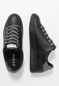 Baskets synthétiques noires avec des accents en paillettes argentées au niveau du talon, semelle texturée et lacets réfléchissants. Étiquette de logo sur la languette.