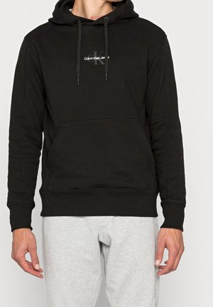 Muž v černé mikině Calvin Klein s přední kapsou a stahovacími šňůrkami, v kombinaci se světle šedými tepláky, zobrazen od krku po boky.