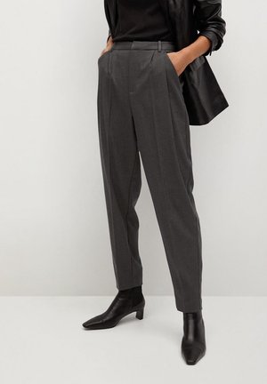 Osoba nosząca szare spodnie z wysokim stanem, czarne skórzane botki za kostkę, czarny top i czarną skórzaną kurtkę z rękami w kieszeniach.