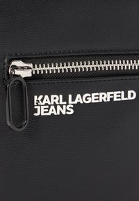 Pelle nera testurizzata con hardware argentato, dotata di una zip e del marchio in rilievo "KARL LAGERFELD JEANS" in bianco.