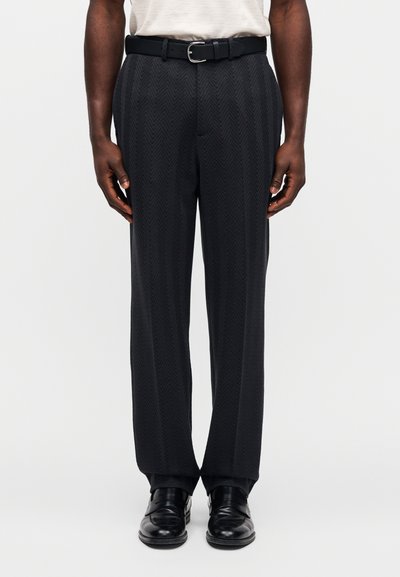 Μαύρο παντελόνι με σχέδιο ψαροκόκαλο και ίσια γραμμή, σε συνδυασμό με μαύρη ζώνη και γυαλιστερά μαύρα παπούτσια. Σχεδιασμός με επίπεδη εμπρόσθια όψη.