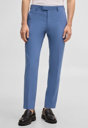 Uomo con pantaloni eleganti blu a taglio slim, mocassini in pelle nera e camicia bianca infilata, su sfondo chiaro e uniforme.