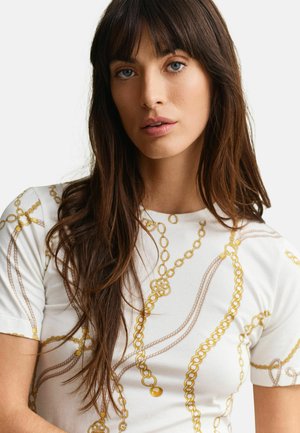 Jonge vrouw met lang bruin haar en blauwe ogen, die een witte blouse draagt met gouden kettingpatronen op een effen achtergrond.