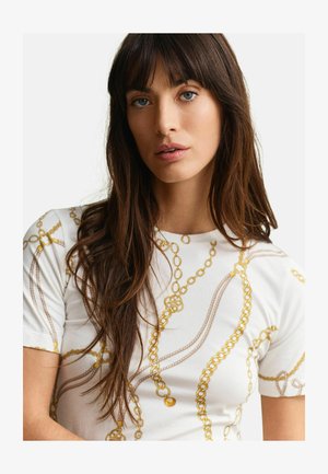 Jeune femme aux longs cheveux bruns et aux yeux bleus portant une chemise blanche ornée de motifs de chaînes dorées sur un fond uni.