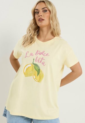 Blondynka w jasnopżółtej koszulce z różowym napisem "La dolce Vita" i grafiką dwóch żółtych cytryn, ubrana w jasnoniebieskie dżinsy.