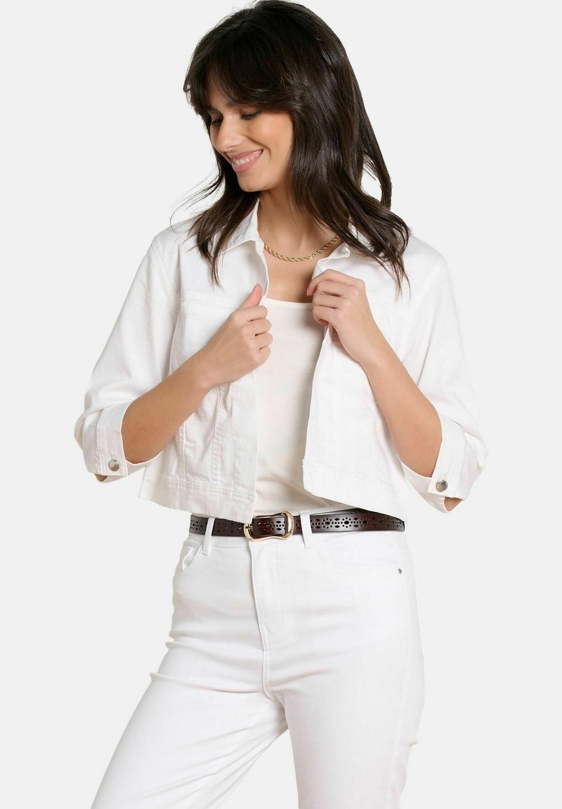 Mujer de cabello oscuro que lleva una chaqueta blanca corta, top blanco, pantalones blancos, un cinturón marrón perforado y un collar de cadena dorada.