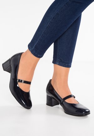 Schwarze Lacklederschuhe mit runder Spitze, einem Knöchelriemen mit Schnalle und einem rechteckigen Blockabsatz. Getragen mit dunkelblauen Jeans.