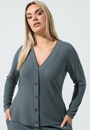 Kvinde med langt blond hår iført en grå ribbet cardigan med knapper og hænderne i lommerne, stående mod en ensfarvet lys baggrund.