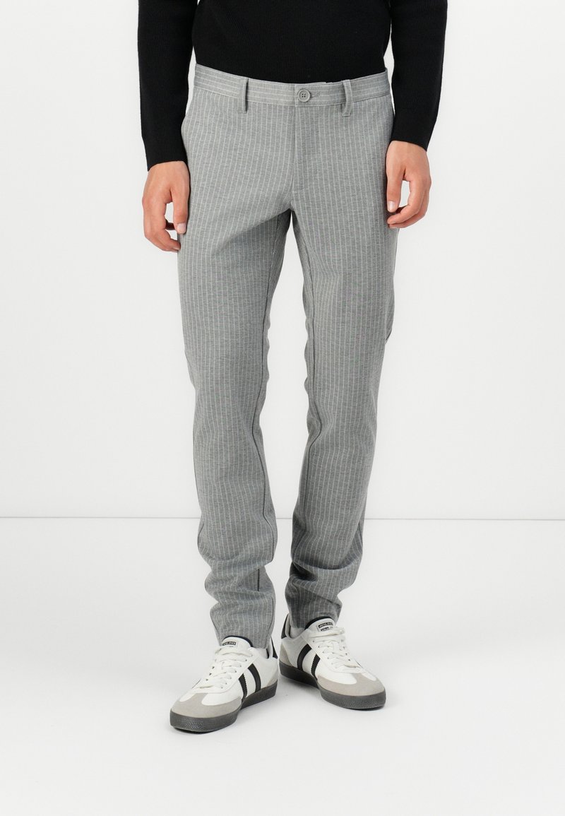Pantalon rayé gris en tissu doux, avec une coupe ajustée, fermeture par boutons et poches. Porté avec des baskets blanches et grises.