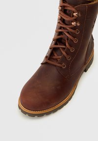 Bota de cuero marrón con cordones que presenta una puntera redondeada, un acabado texturizado, ojales de metal y una suela de goma con un borde en contraste de color marrón claro.
