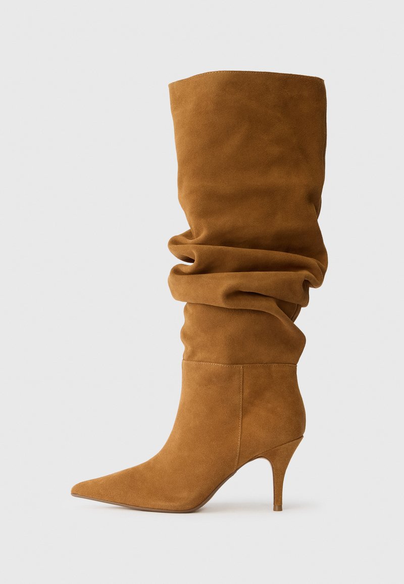 Bottines en daim marron, montant jusqu'au genou, avec un design décontracté, un bout pointu et un talon aiguille, présentant une texture douce et un bord supérieur lisse.