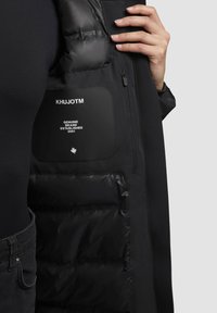Nahaufnahme einer Hand, die die schwarze gepolsterte Jacke aufhält und eine Innentasche mit dem Text "KHUJOTM Genuine Brand Established 2001" zeigt.