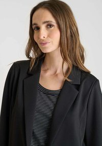 Blazer noir au design structuré, avec un col cranté. Porté par-dessus un haut à rayures noir et blanc, en tissu lisse et avec une coupe ajustée.