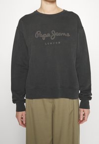 Sudadera gris oscuro con cuello redondo, que presenta un logotipo brillante de "Pepe Jeans London". Mangas largas y corte relajado. El material parece suave.