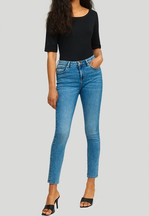 Nainen pukeutuneena istuvaan mustaan toppiin, korkeavyötäröisiin sinisiin skinny-farkkuihin ja mustiin avokkaisiin korkokengillä, seisoo yhdellä kädellä taskussa.