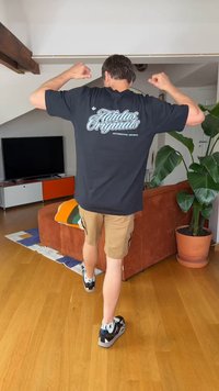 Homme portant un t-shirt noir Adidas Originals et un short beige lève les bras dans un salon avec canapé, télévision et grande plante en pot.