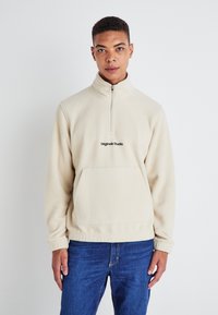 Beige fleece sweatshirt with a half-zip collar, front pocket, and black logo on the chest. Soft texture, relaxed fit, and long sleeves.