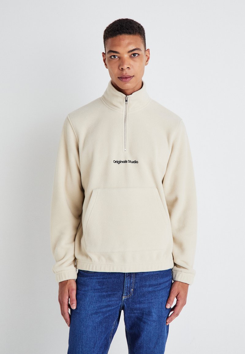 Beige fleece sweatshirt with a half-zip collar, front pocket, and black logo on the chest. Soft texture, relaxed fit, and long sleeves.