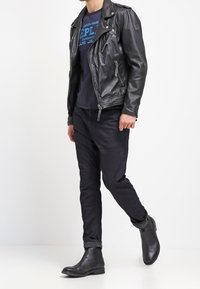 Giacca da motociclista in pelle nera, t-shirt grafica blu, pantaloni scuri e stivaletti neri. Materiali texturizzati e design aderente.