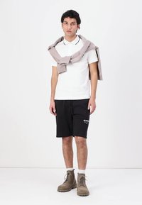Hvit polo skjorte med svarte detaljer, svarte shorts med logo, beige genser slengt over skuldrene, beige støvler, stående mot en ensfarget bakgrunn.