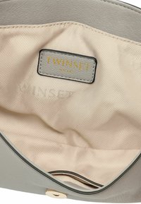Interno di una borsa in pelle grigia con una fodera beige a righe e un'etichetta del marchio stampata in oro con scritto "TWINSET". Dettaglio della chiusura magnetica visibile.