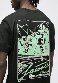 Juoda marškinėliai su mėtų žalia kalnų ir medžių grafika, apatiniame kampe pavaizduotas teniso kortas ir užrašas „Lacoste Tennis Heritage“.