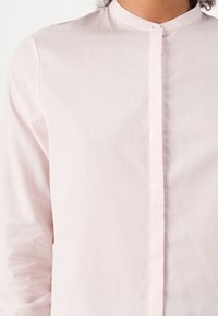Camicia button-up rosa chiaro con colletto alla cinese, realizzata in tessuto morbido, con una placca frontale e maniche lunghe.