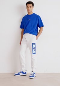 Ein blaues T-Shirt mit Rundhalsausschnitt, kombiniert mit weißen Jogginghosen, die ein blaues "HUGO"-Logo tragen. Sneakers mit blauen und weißen Akzenten runden das Outfit ab.