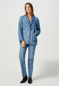 Vaaleansininen denim-takkimallinen bleiseri, jossa on lovellinen kaulus, kaksi etupocketsia ja kaksi nappia; yhdistettynä vastaaviin denim-housuihin ja mustiin nilkkabootseihin.