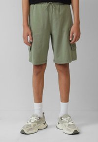 Olivgrüne Cargo-Shorts aus leichtem Material mit einem elastischen Bund, ausgestattet mit Seitentaschen und einem lässigen Schnitt. Getragen mit grauen Turnschuhen.