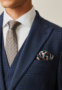 Chaqueta de traje de cuadros en azul marino con solapas de pico, una corbata beige con patrones azules y un colorido pañuelo de seda en el bolsillo con un diseño geométrico.